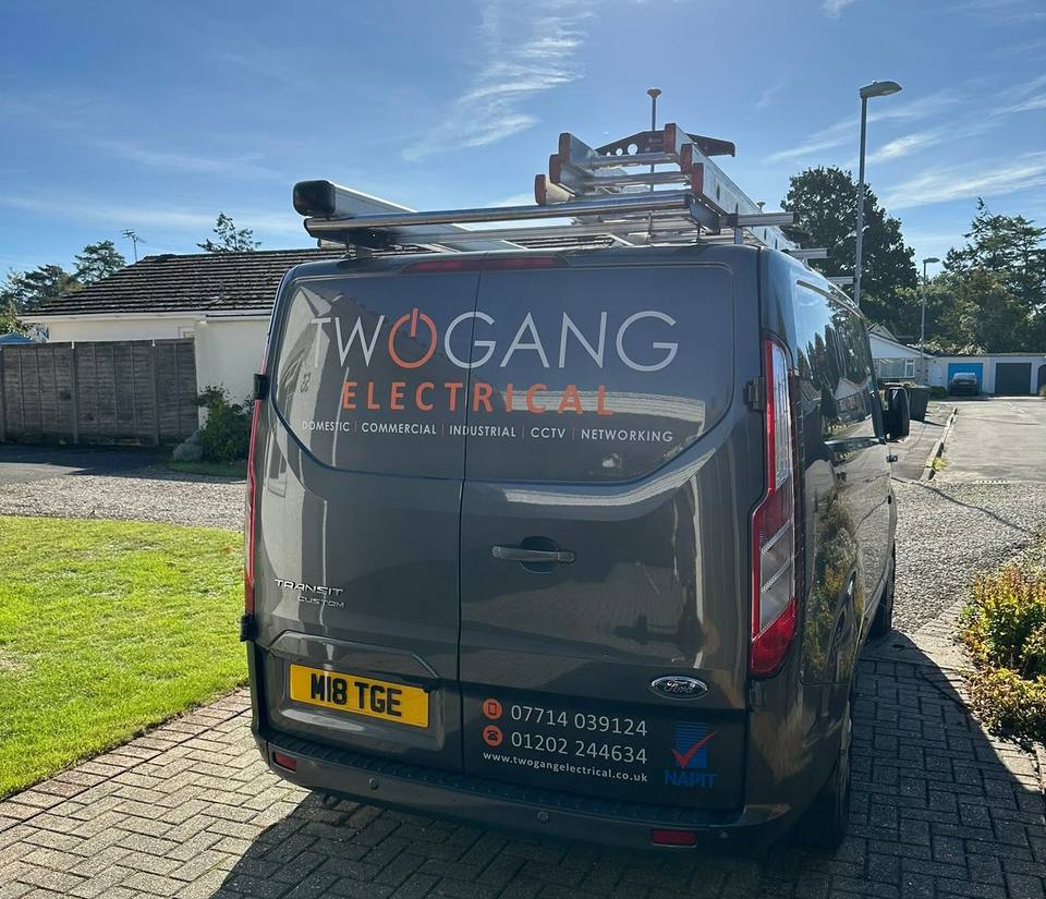A sign-written Two Gang Electrical van with a ladder on the roof rack, parked on a block-paved driveway