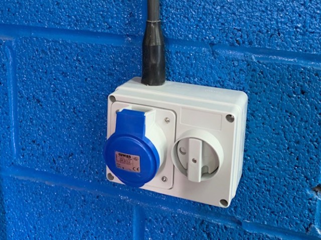 A blue single-phase industrial socket with an integrated isolator, surface-mounted on a painted block wall